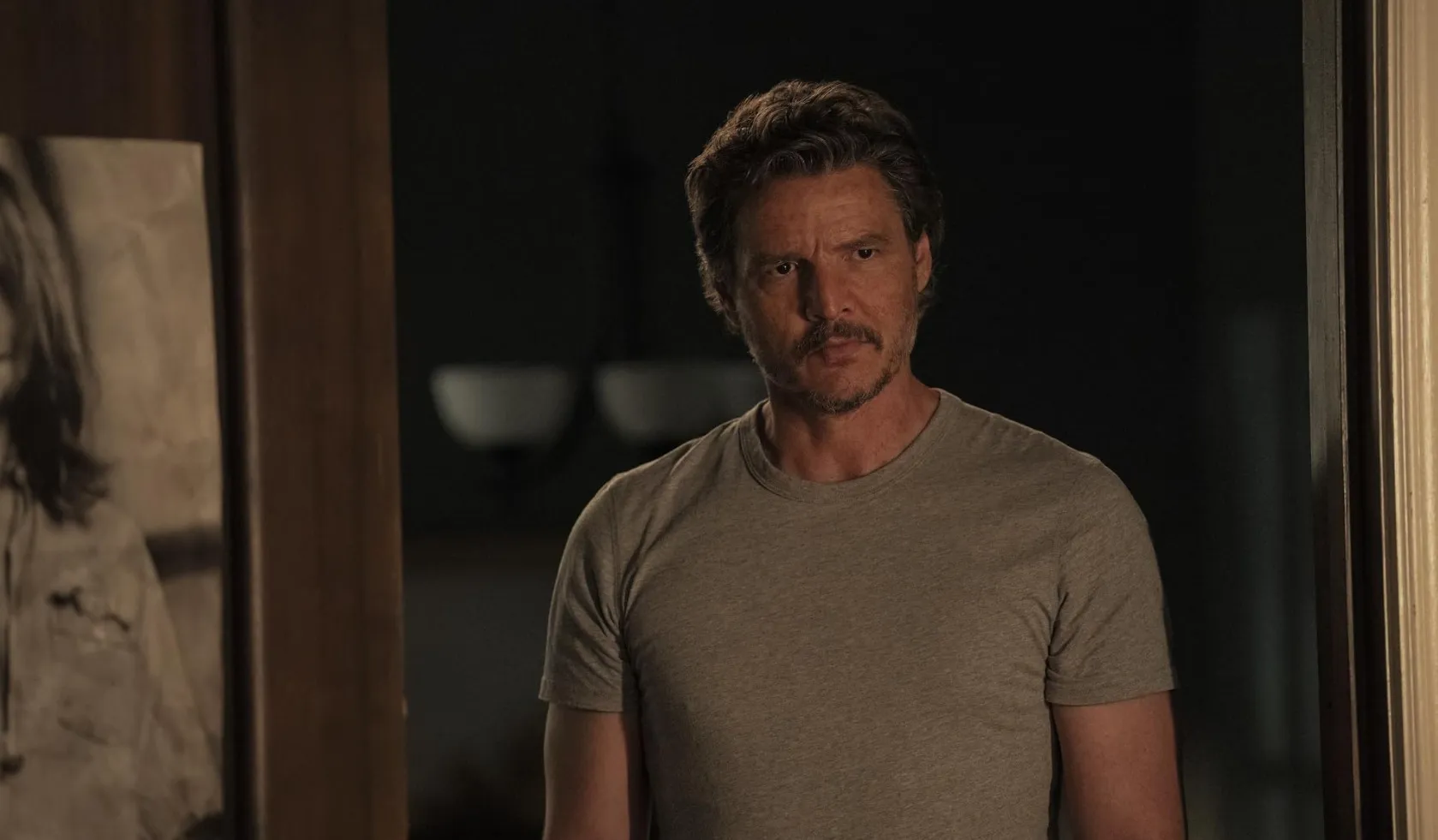 Man in grey t-shirt with mustache and beard standing indoors, illustrating actors A-Z in a serious mood.