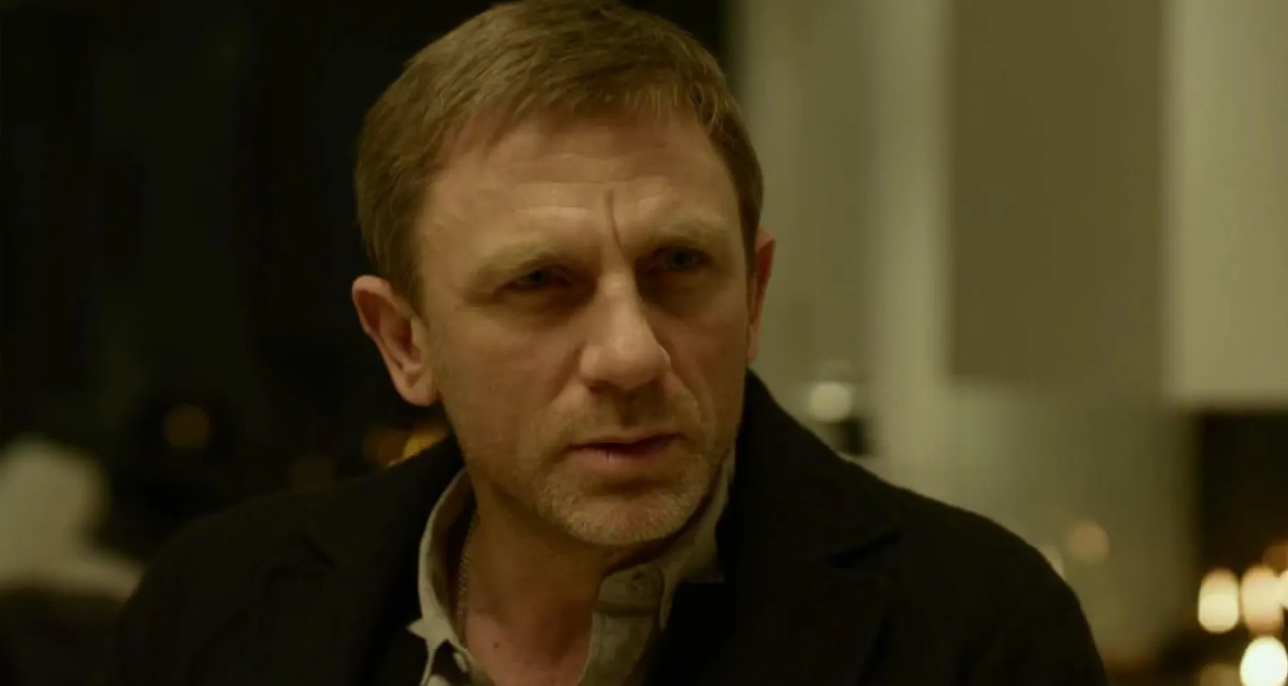 Daniel Craig in a close-up scene from a film, showing a serious expression in a low-lit indoor setting.