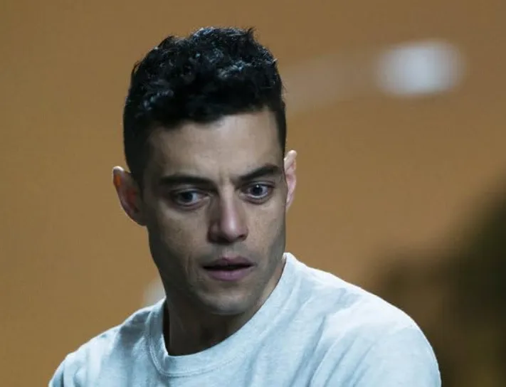 Rami Malek portraying Freddie Mercury with intense expression, wearing a light gray shirt against a blurred background