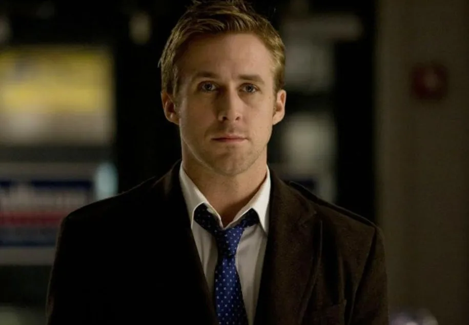 Ryan Gosling in a dark suit and blue tie, looking directly at the camera in a dimly lit setting.