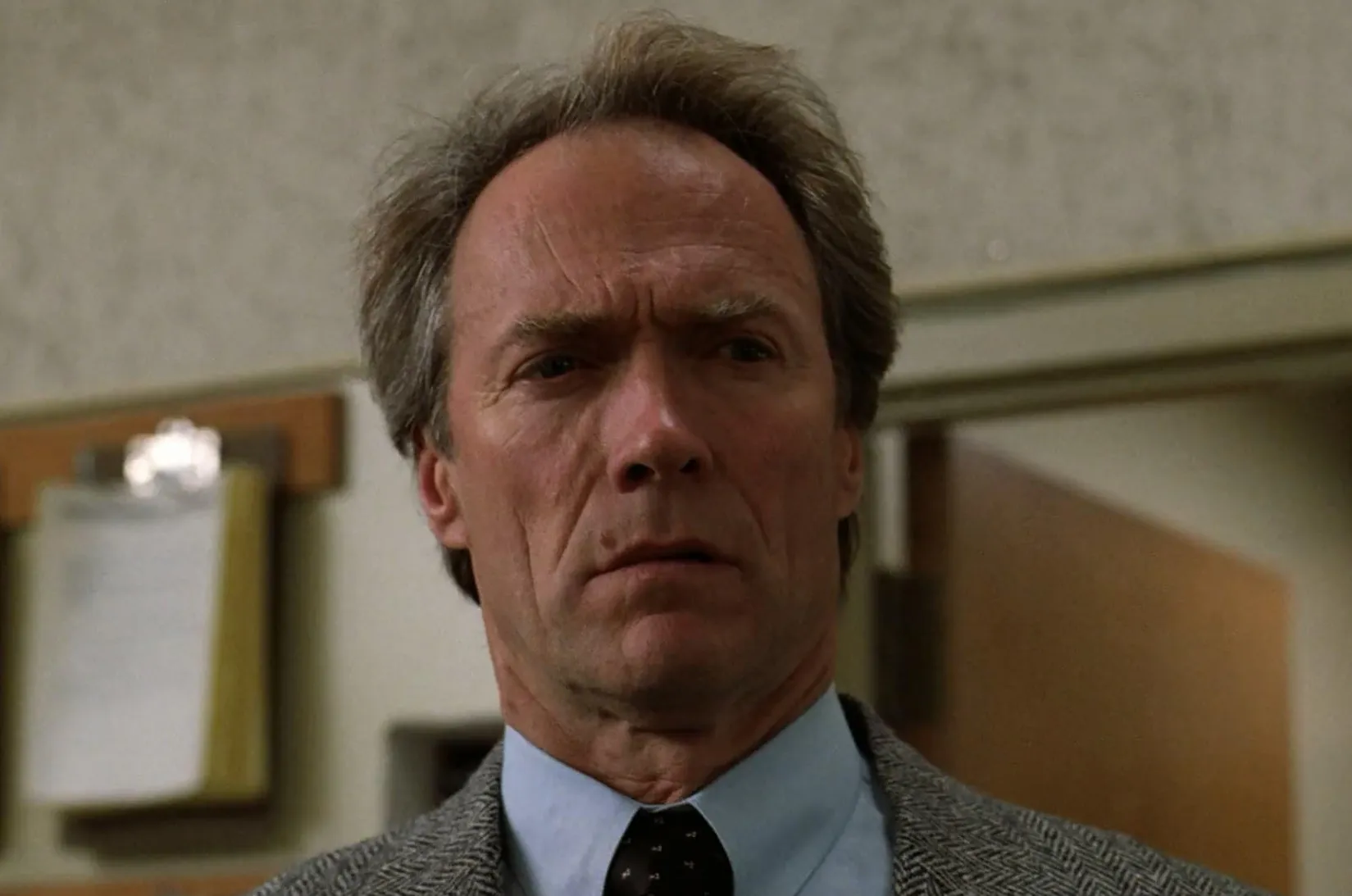 Clint Eastwood in a serious expression, wearing a suit and tie, in an indoor setting from a film scene.