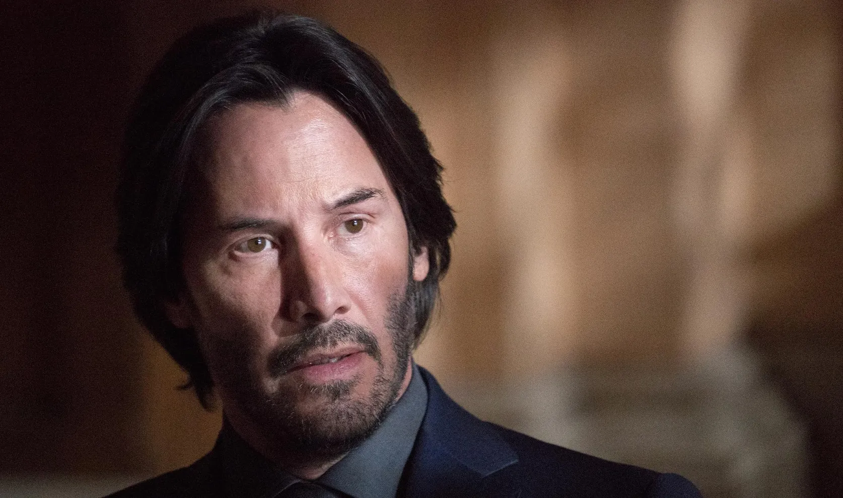Keanu Reeves in a close-up scene from a film ending in X, dressed in dark suit with serious expression.