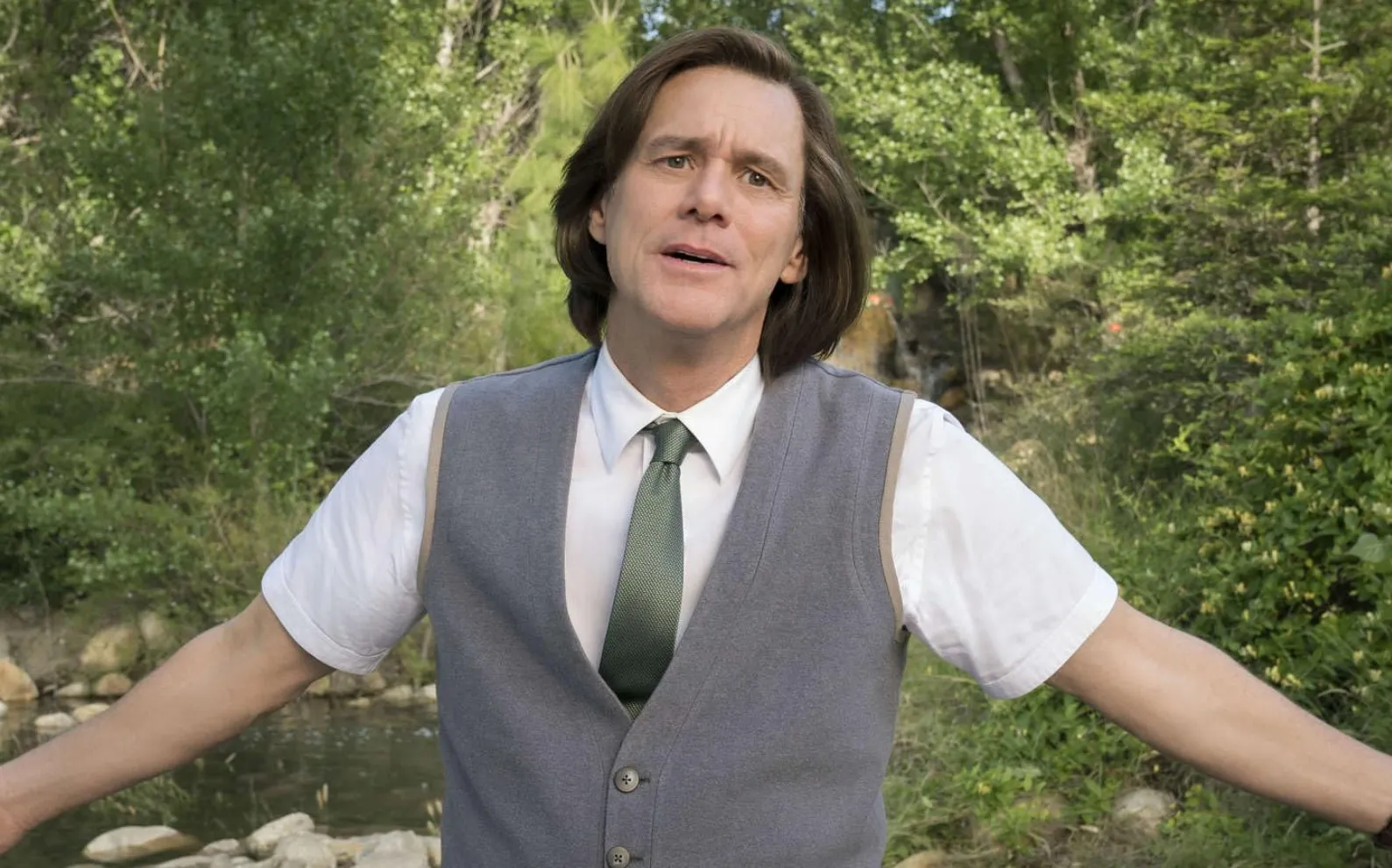 Jim Carrey in a gray vest, white shirt, and green tie standing outside near a creek with trees in the background.
