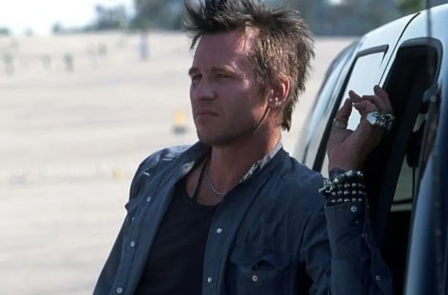 Actor Val Kilmer leaning against a vehicle wearing a dark shirt with spiked bracelets in an outdoor scene.