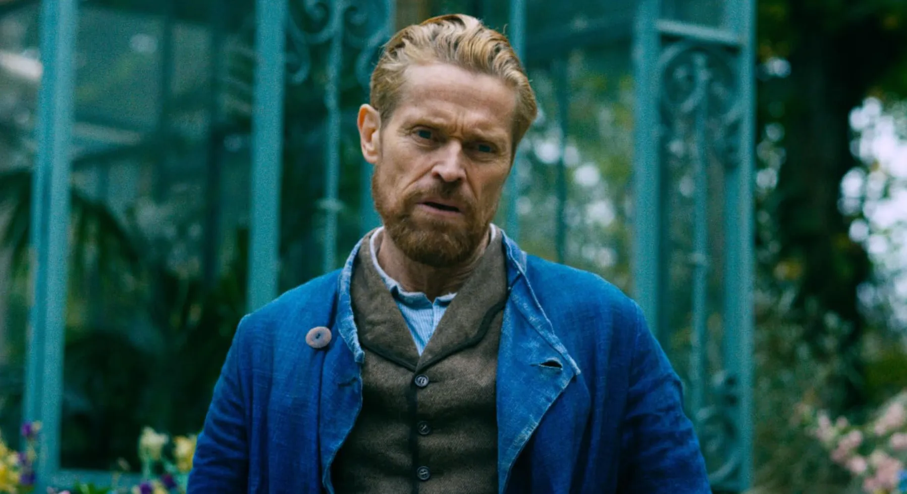 Male actor with blond hair and beard wearing a blue coat and brown vest in an outdoor setting with plants and glass.