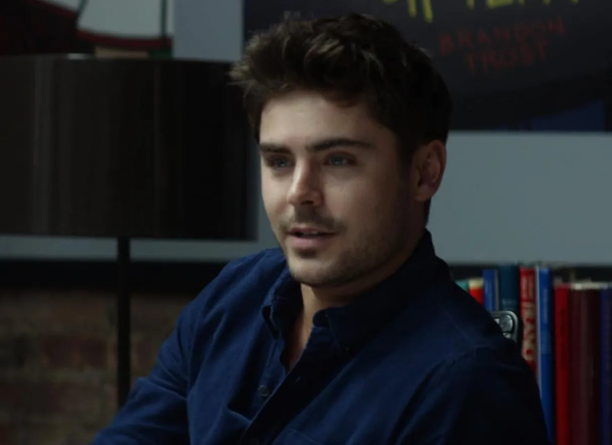 Zac Efron wearing a dark blue shirt sitting indoors with books and a lamp in the background.