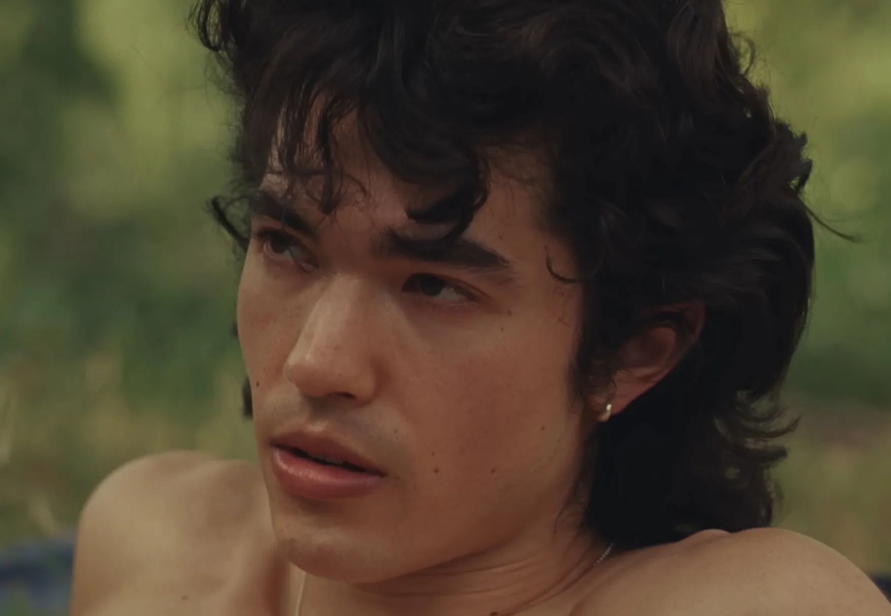 Close-up of celebrity Conan Gray making eye contact, outdoors with natural green background and tousled dark hair.