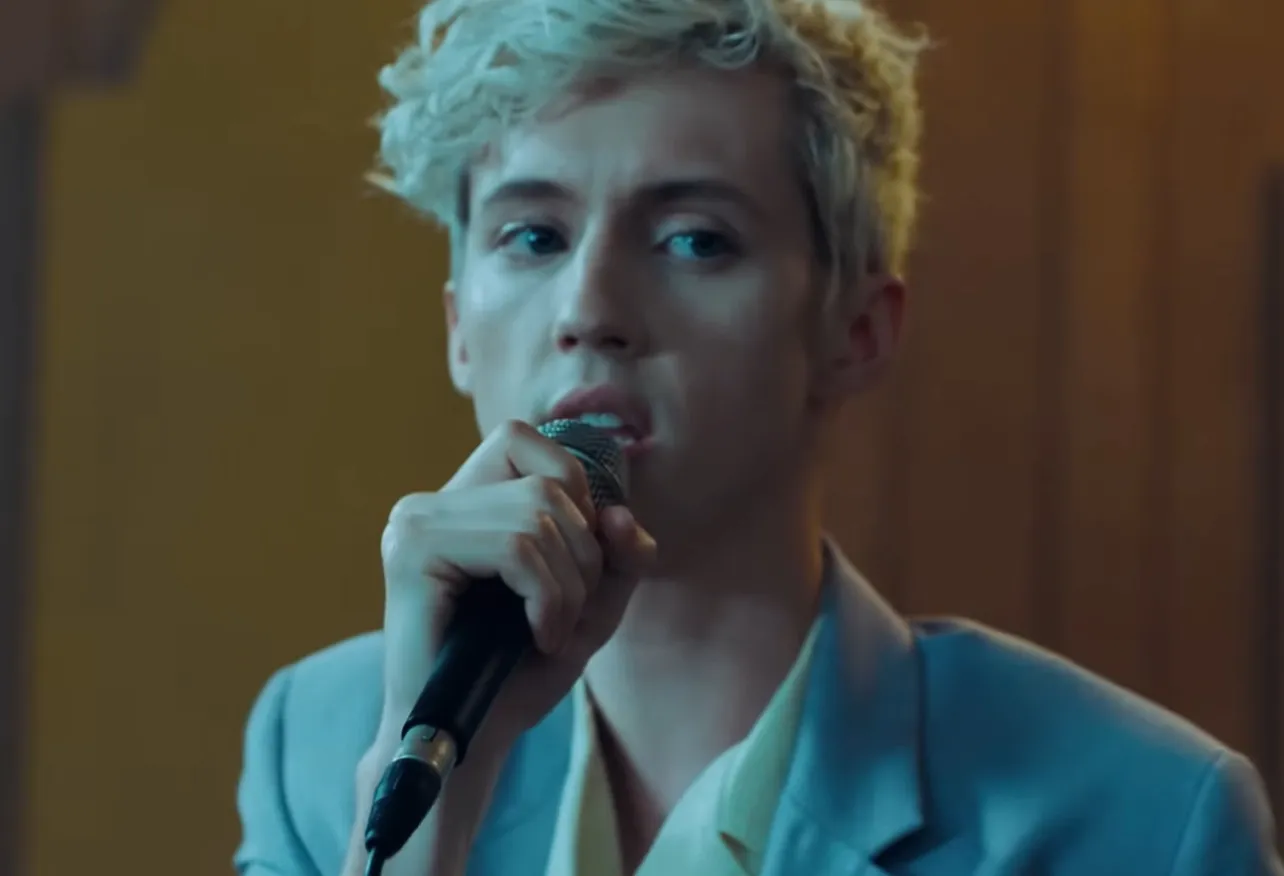 Troye Sivan making eye contact while singing into a microphone, wearing a light blue blazer in a warm setting