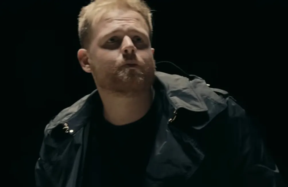 Tom Walker with intense eyes, wearing a dark jacket against a black background in a low-light setting