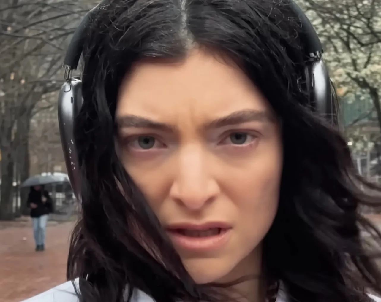 Close-up of Lorde's eyes with dark hair and headphones outdoors on a cloudy day with blurred background trees.