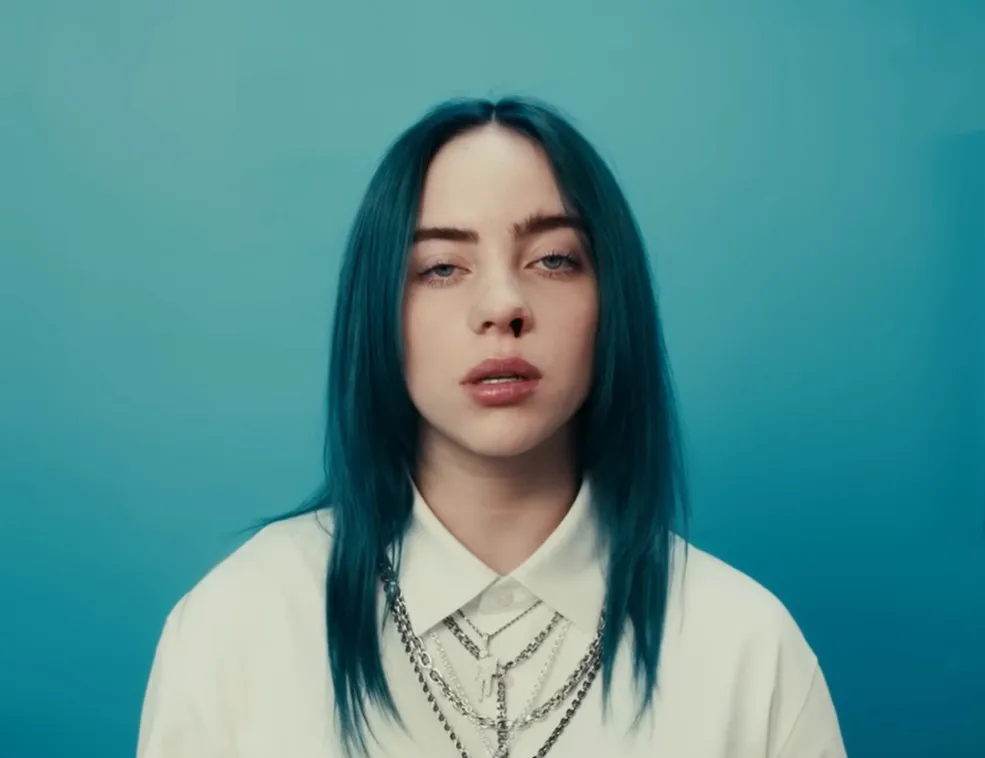 Billie Eilish with teal hair and layered silver chains wearing a white collared shirt against a blue background