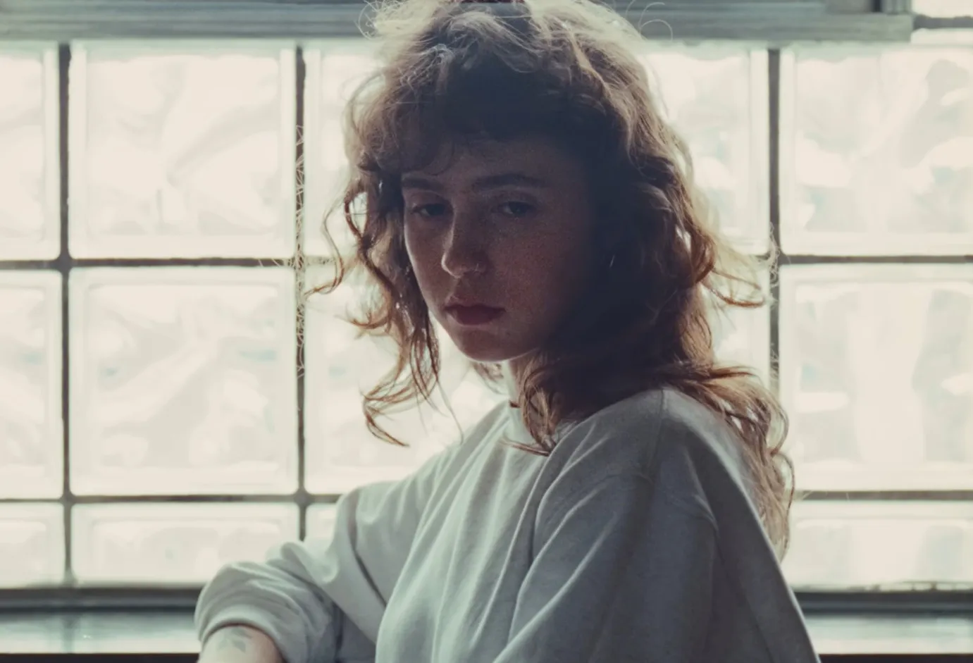 Young woman with curly hair and intense eyes sitting near a frosted window, focusing on celebrity eyes Clairo.