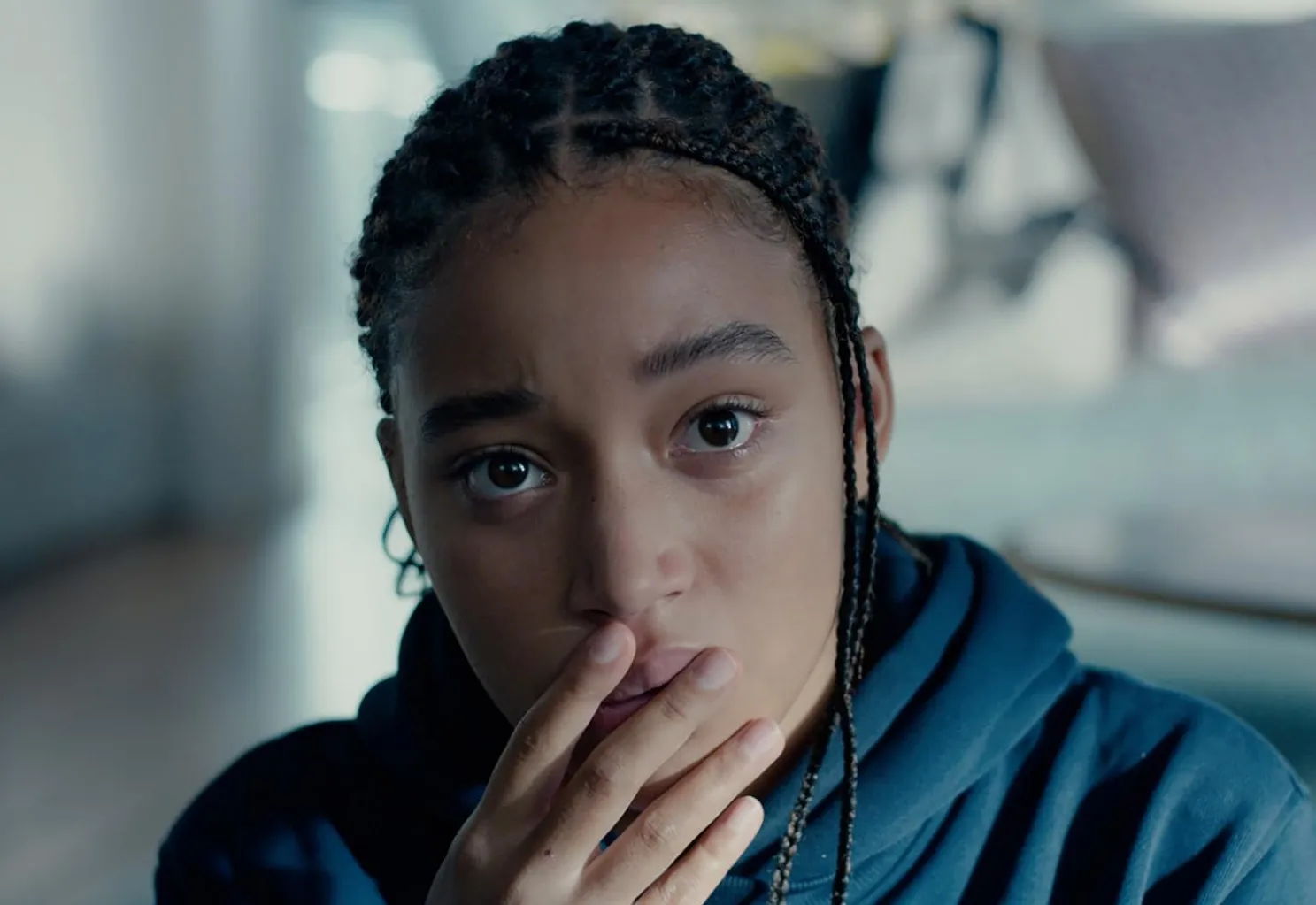 Amandla Stenberg looking contemplative with braided hair and hand partially covering mouth in a casual setting.