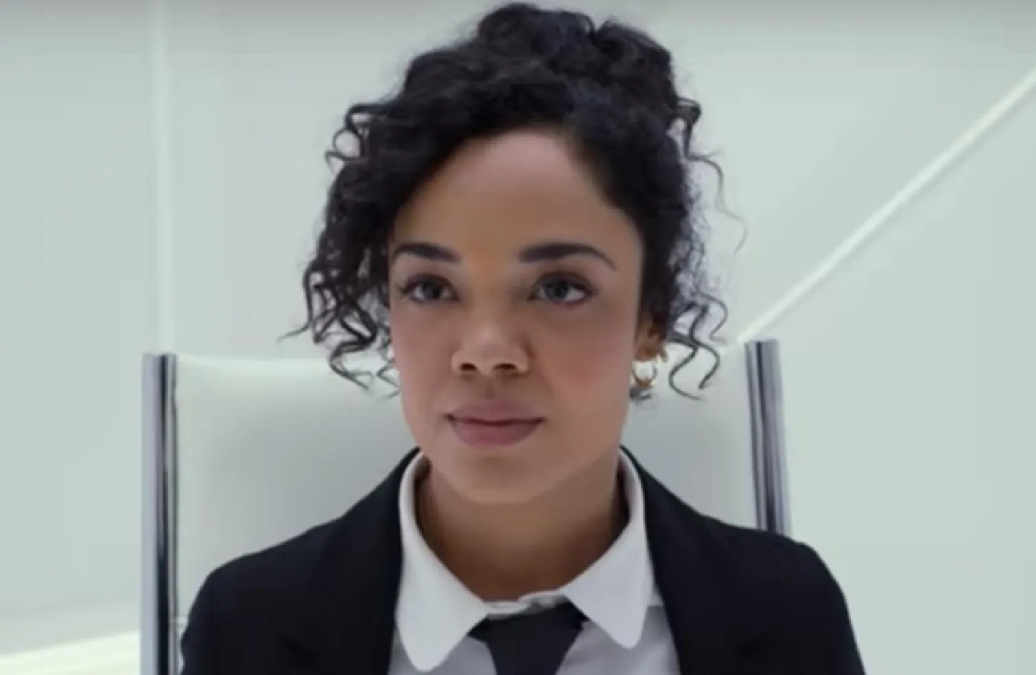 Tessa Thompson looking off into the distance, wearing a black suit and white shirt in a bright room.