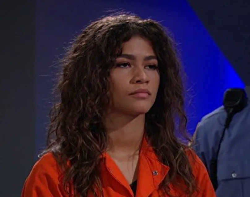 Zendaya with curly hair wearing an orange outfit, looking off into the distance in a dimly lit setting.