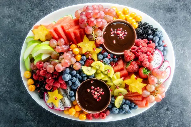 Colorful store-bought fruit platter with grapes, kiwi, berries, starfruit, watermelon, and chocolate dipping sauce on a white plate.