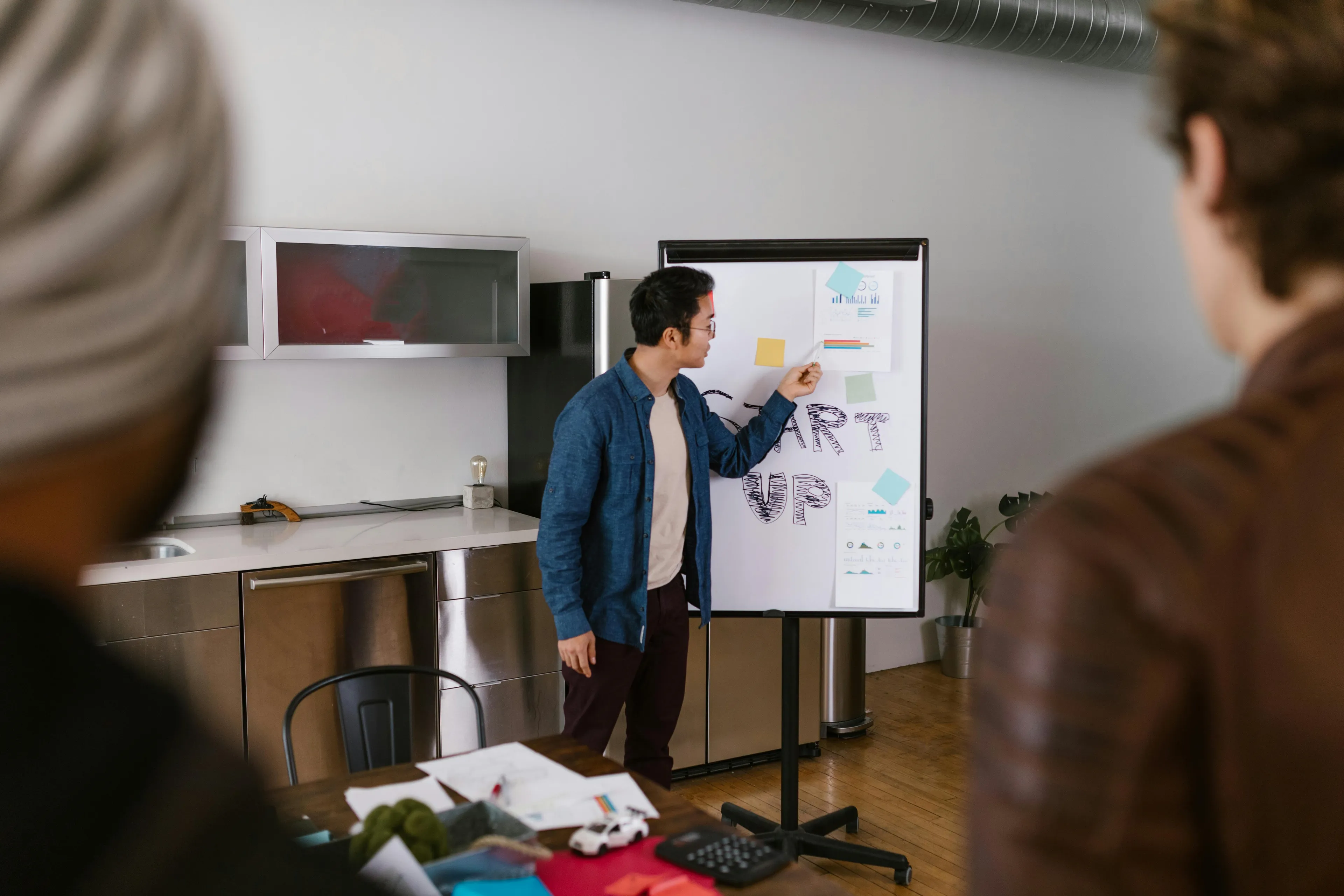 Man preparing presentation at office, pointing to notes on flip chart while two colleagues observe attentively.