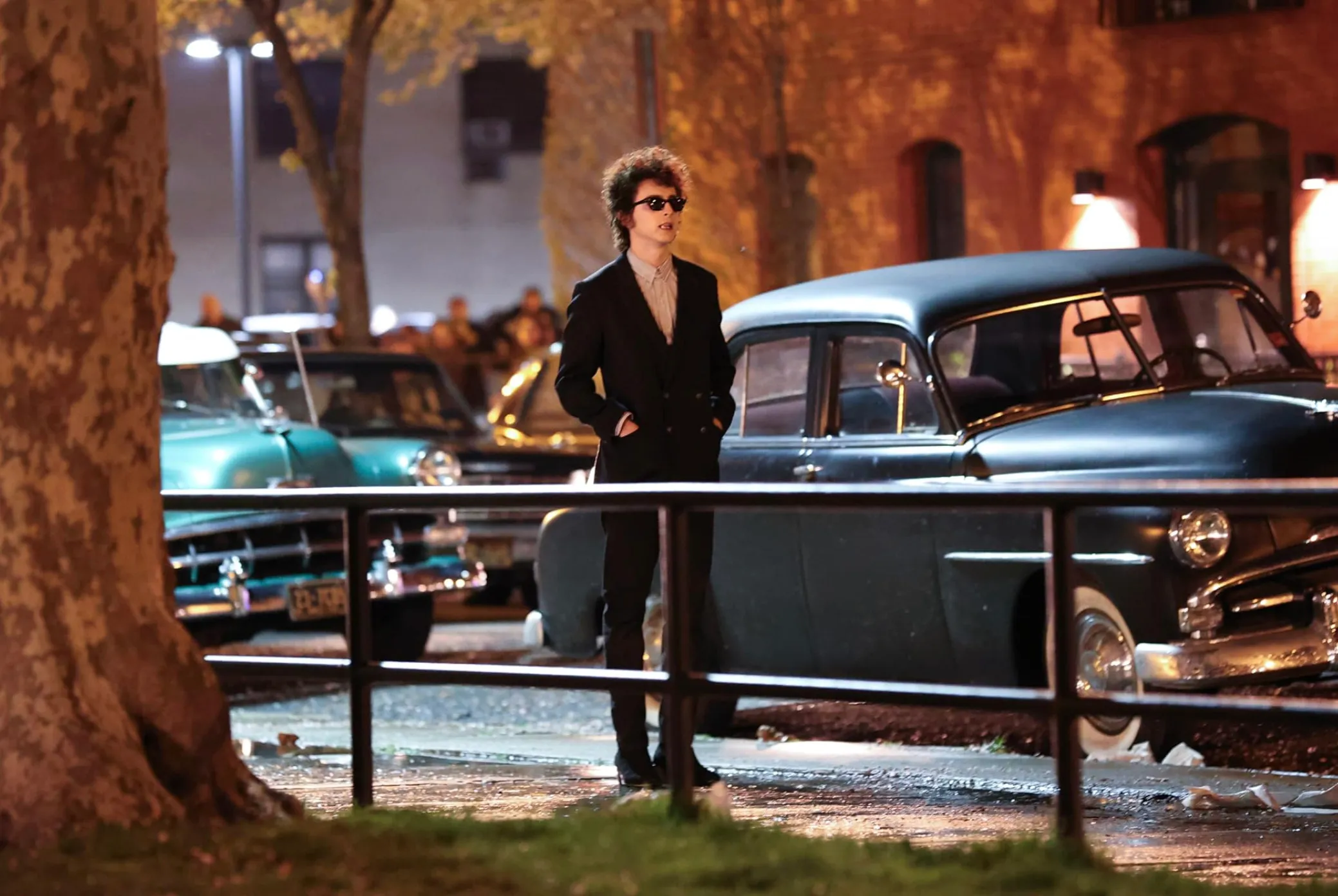 Timothée Chalamet in A Complete Unknown movie scene, standing near classic cars at night in urban setting.