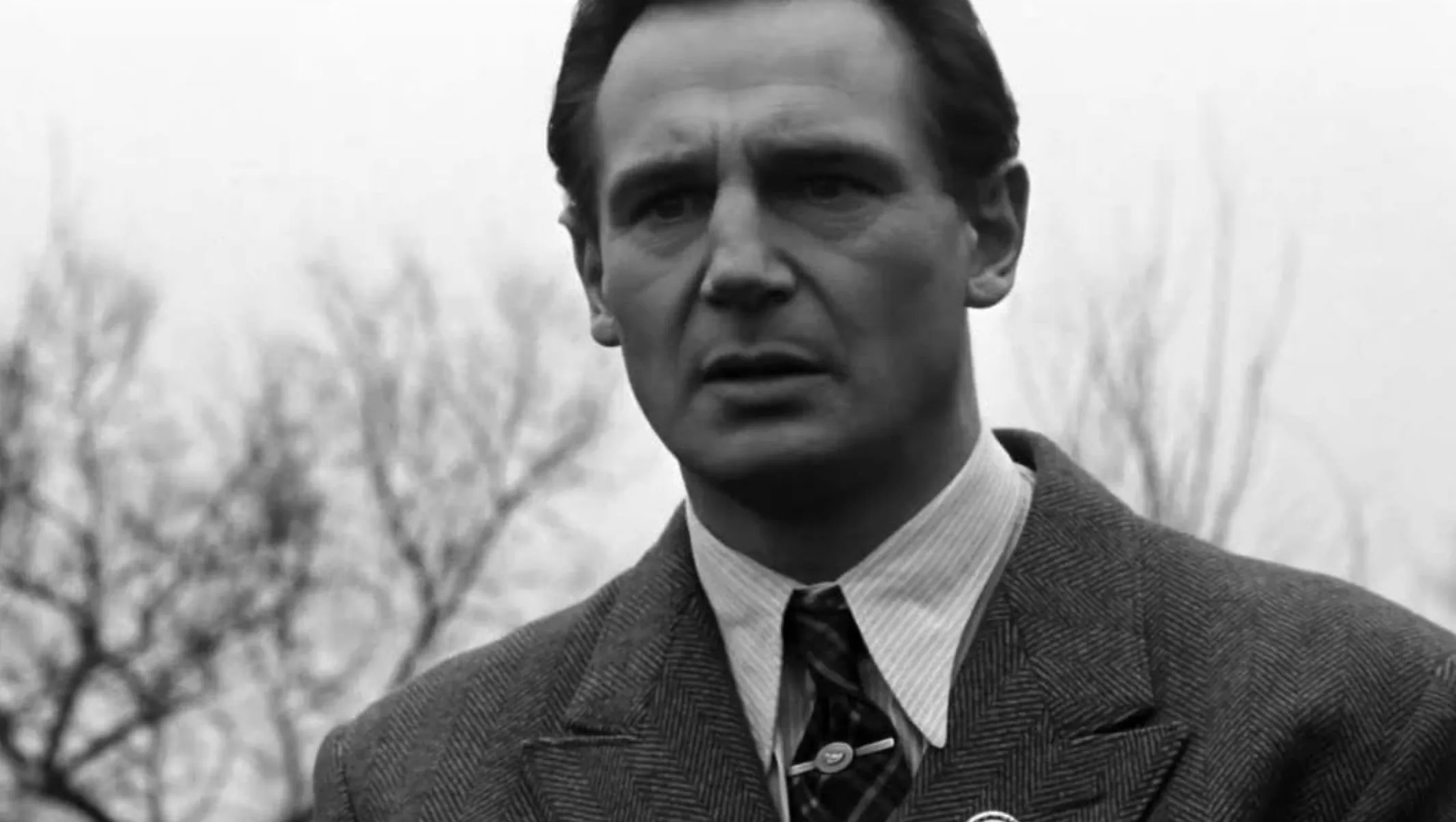 Liam Neeson on screen portraying a serious character known for intense and dramatic roles in a black and white movie scene.