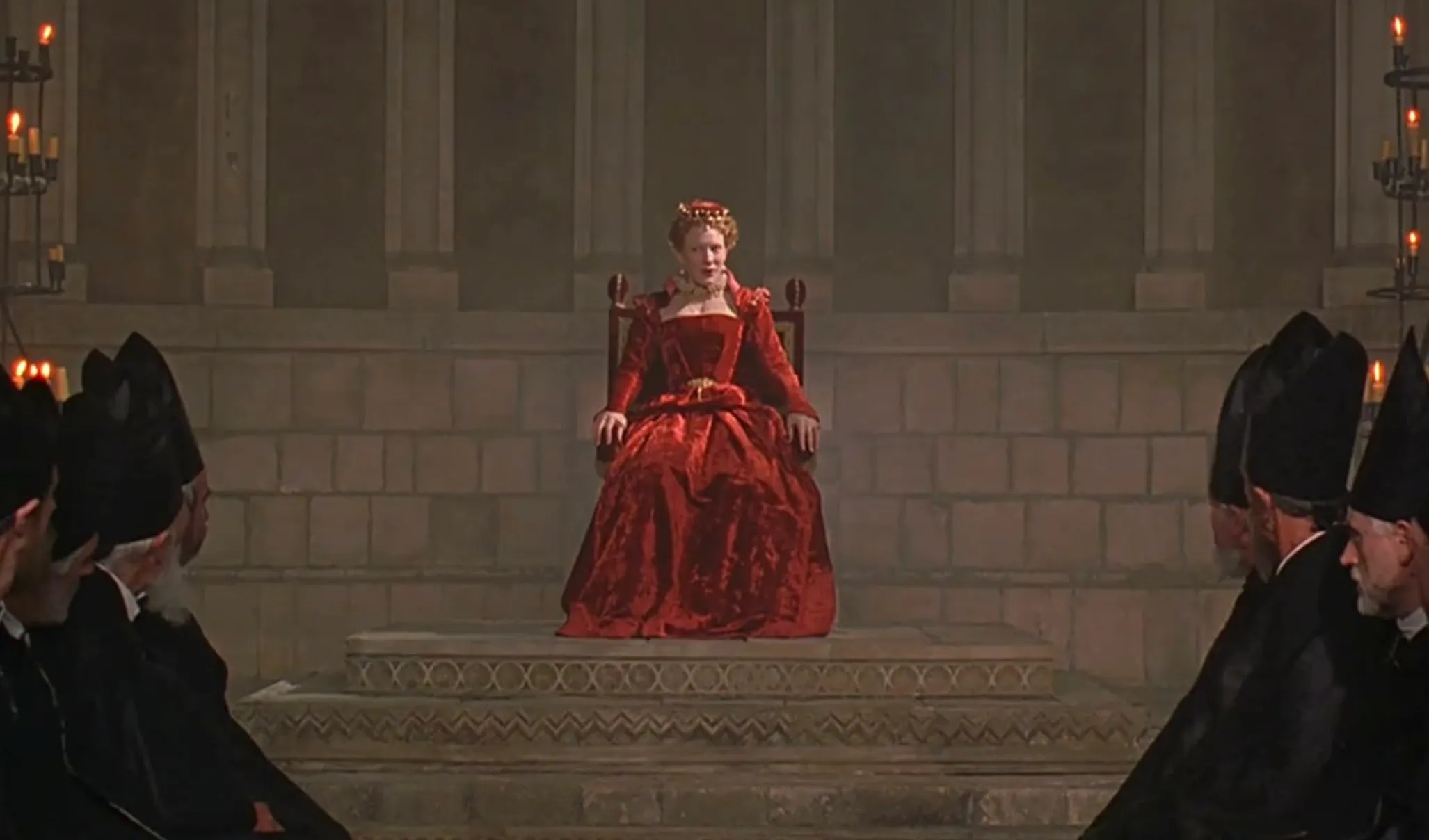 Cate Blanchett portraying Queen Elizabeth I in a 1998 movie scene seated on a throne in a grand hall.