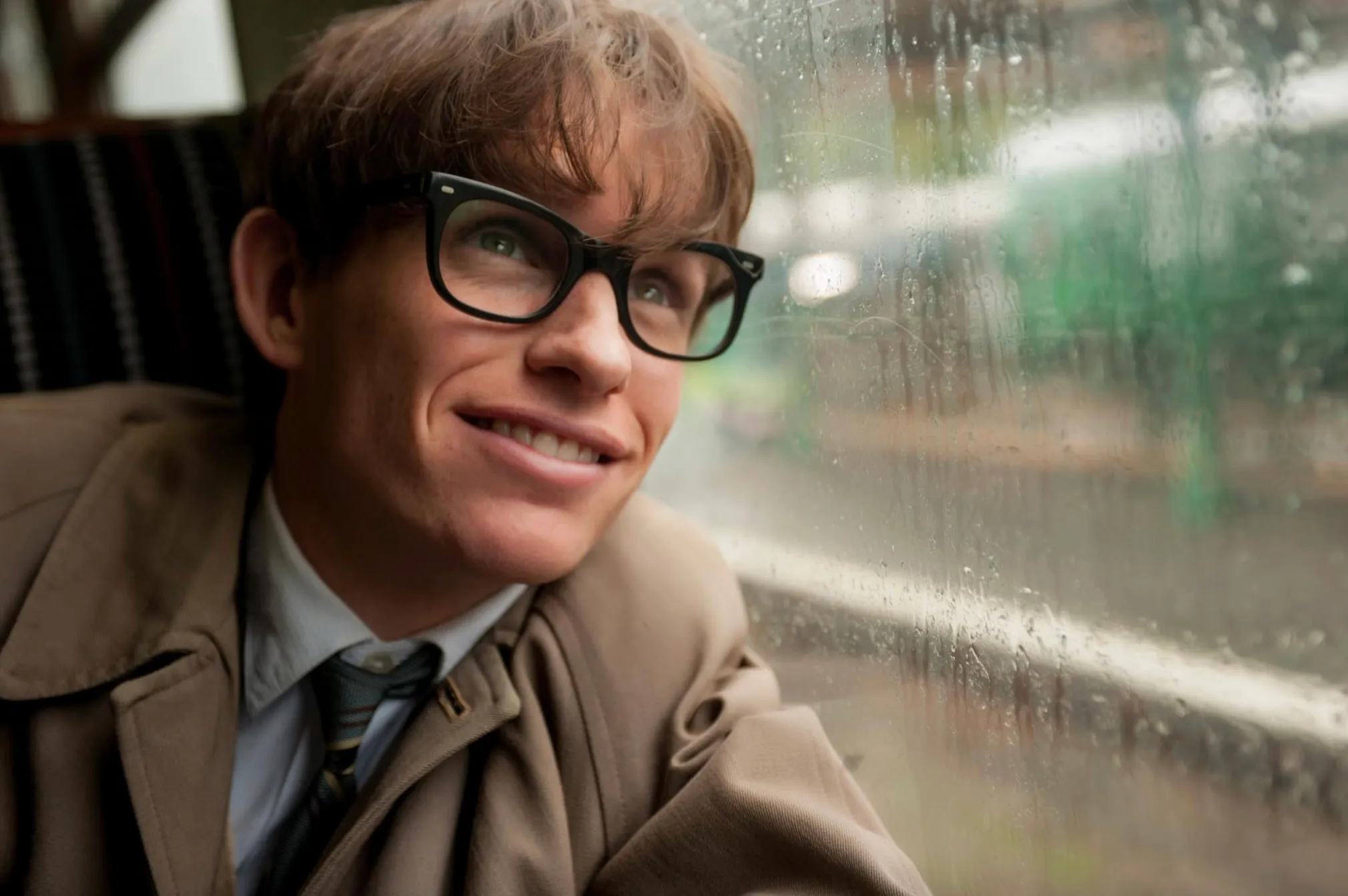Eddie Redmayne as Stephen Hawking in The Theory of Everything, smiling and looking out a rainy window.