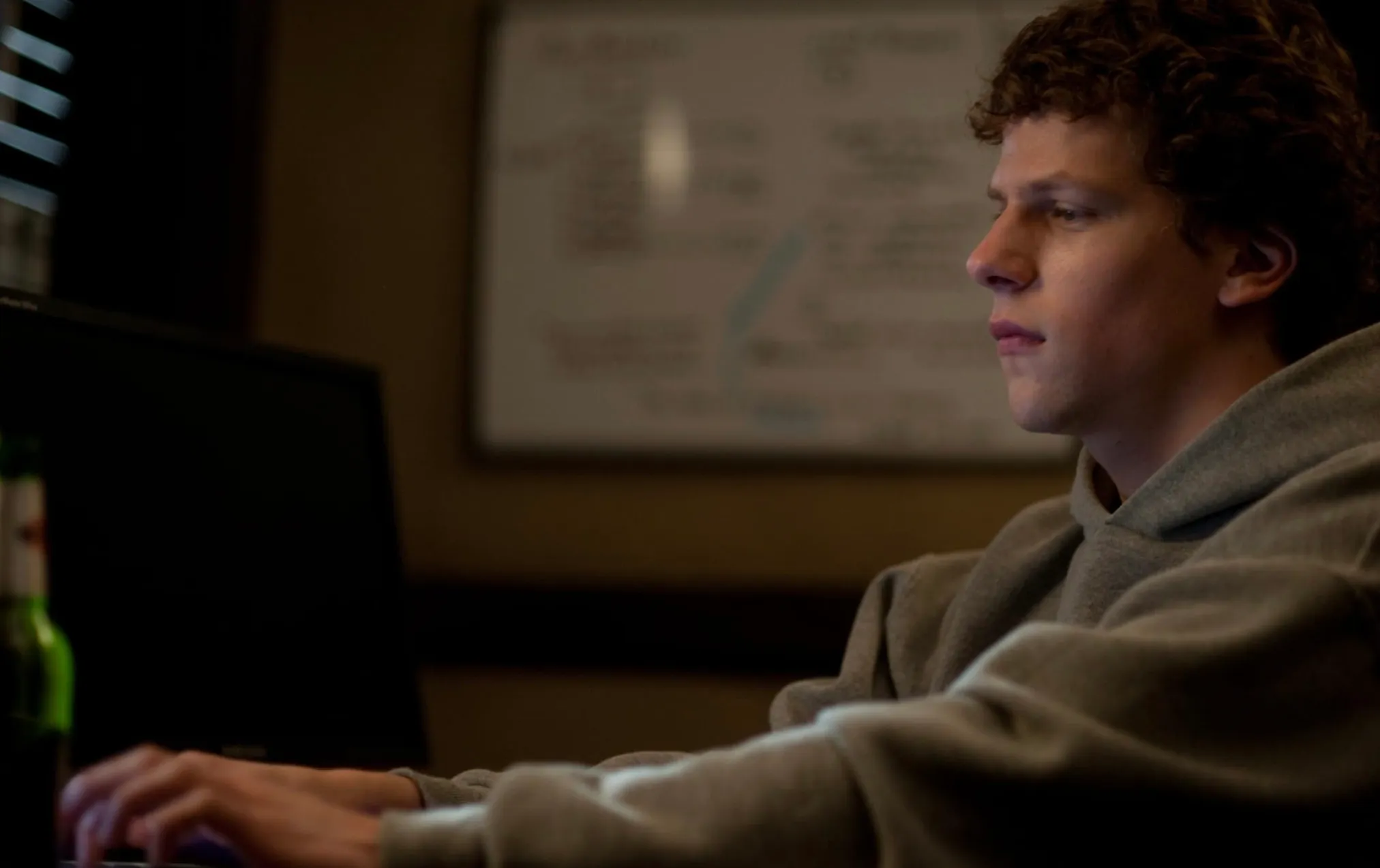 Jesse Eisenberg portraying Mark Zuckerberg in The Social Network, focused on a computer screen in dim lighting.