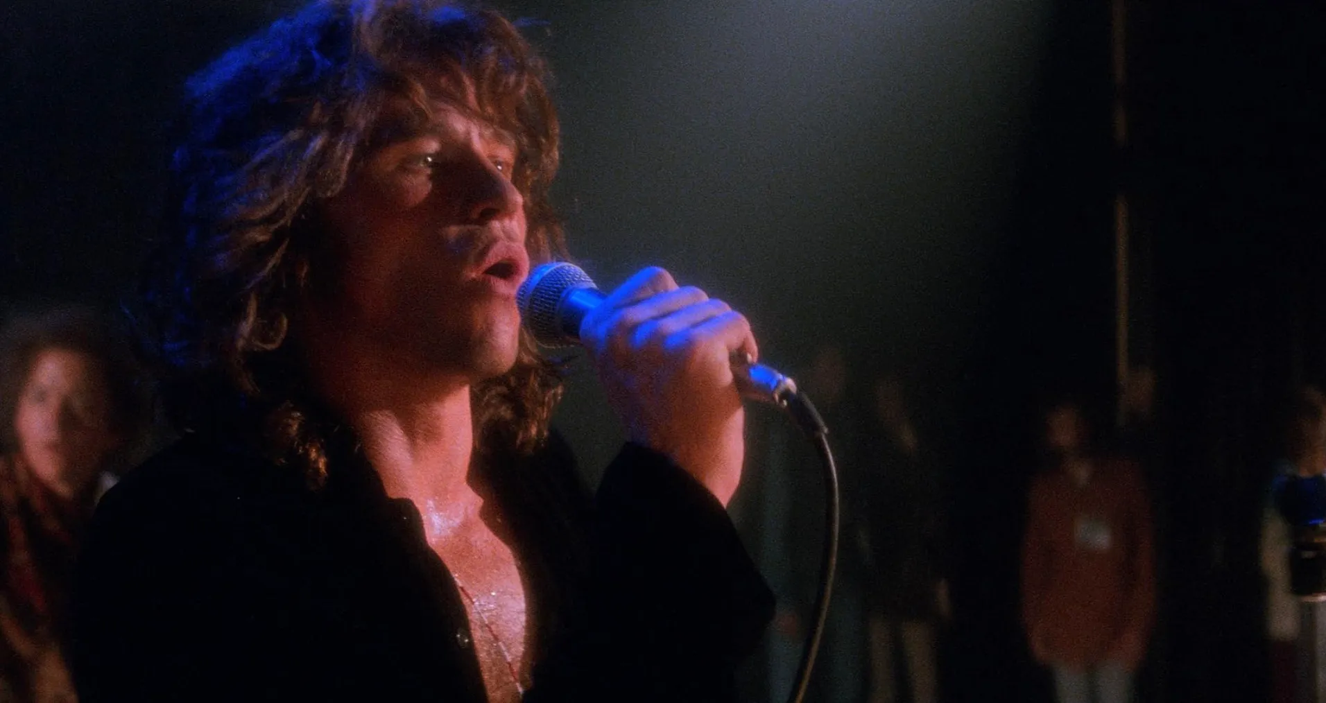 Actor singing into a microphone on stage, famous people on screens, who did this actor portray in a dramatic scene.