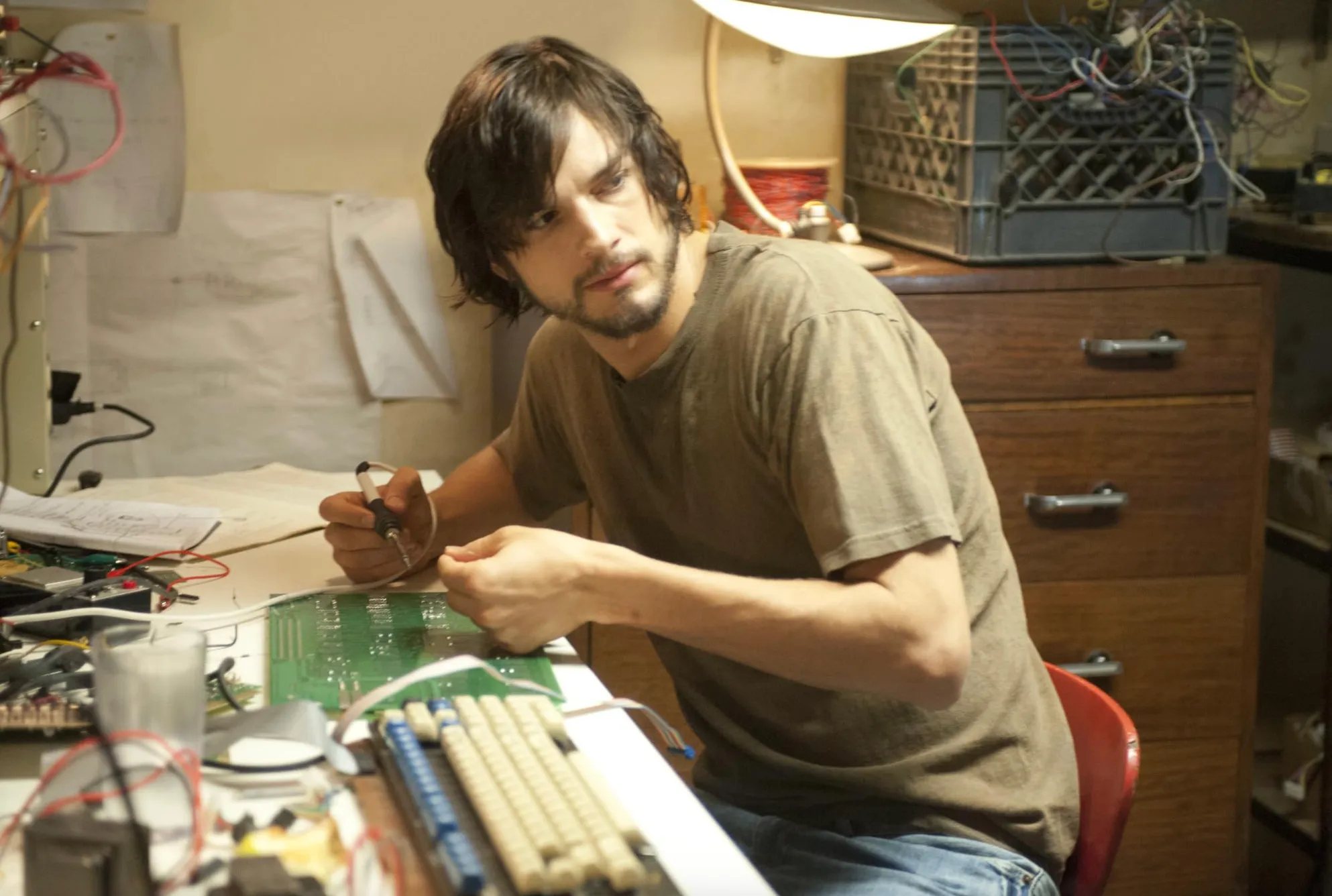 Ashton Kutcher as a famous character working on electronics at a cluttered desk in a dimly lit room.