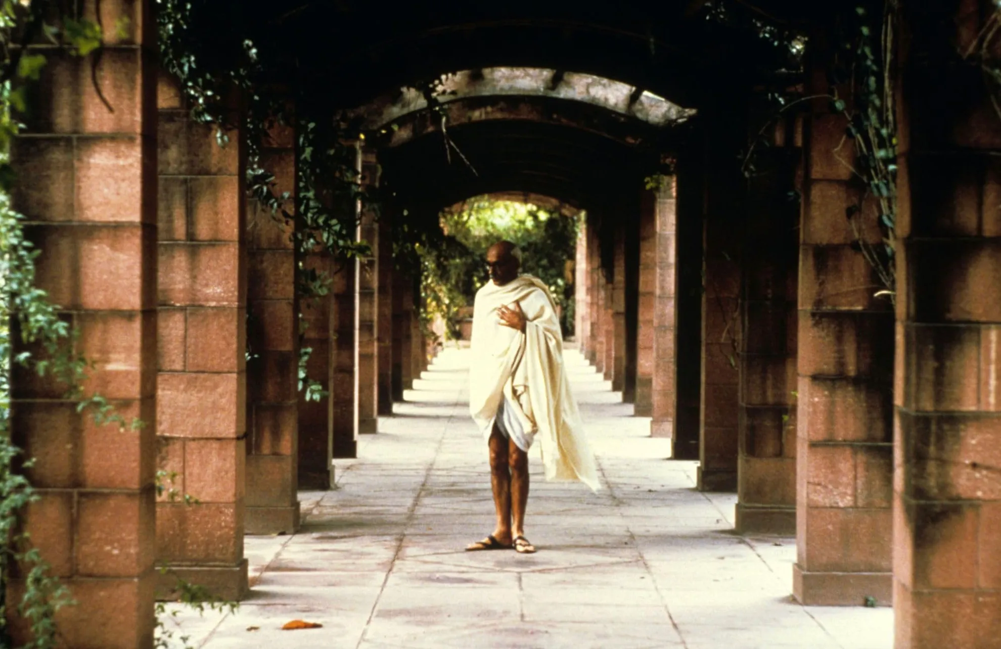 Ben Kingsley portraying Mahatma Gandhi in the 1982 movie walking through an arched stone corridor.