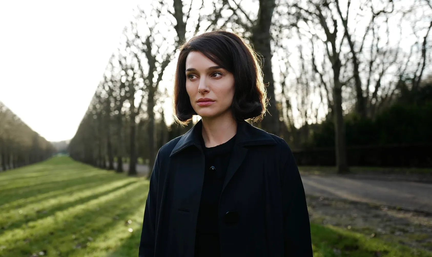 Natalie Portman portraying a character in the historical drama film Jackie, standing outdoors in a dark coat.