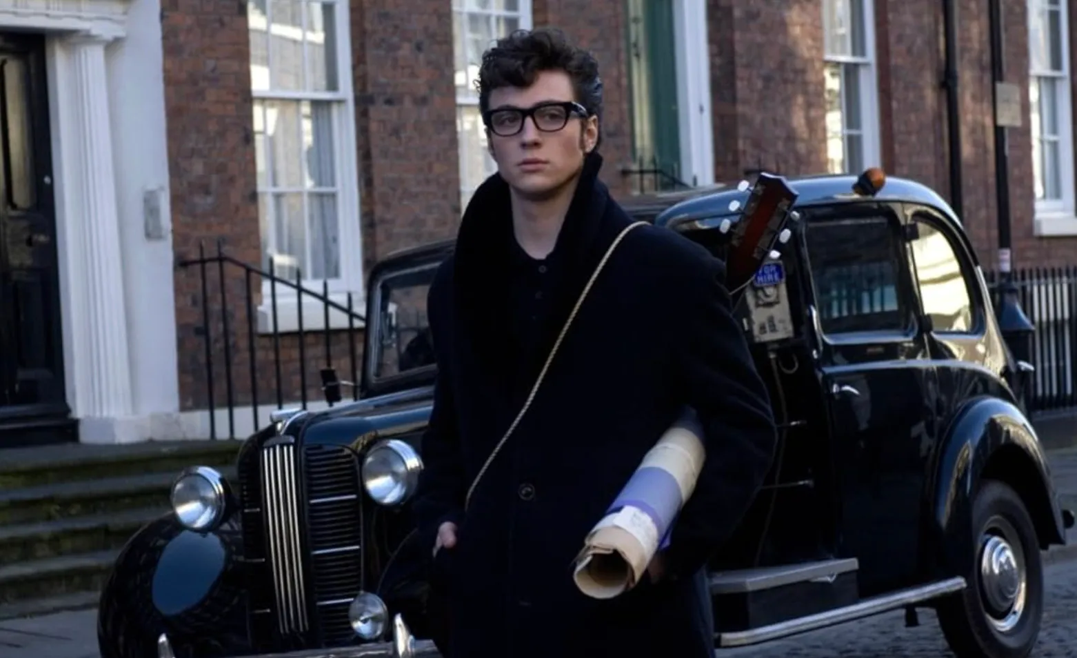 Young man resembling Aaron Perry Taylor-Johnson standing by a vintage car in a scene from a 2009 movie portrait.