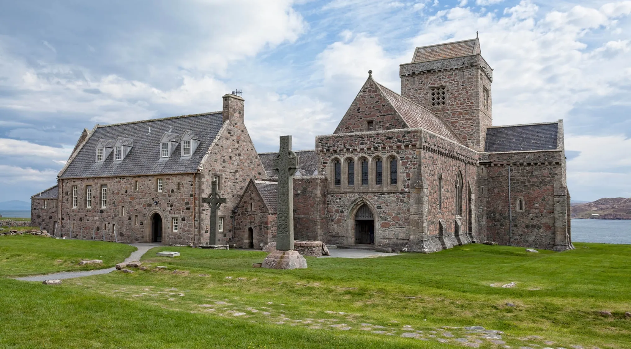 Historic stone Church of Scotland building with Celtic cross on green grass under a cloudy sky by the sea.