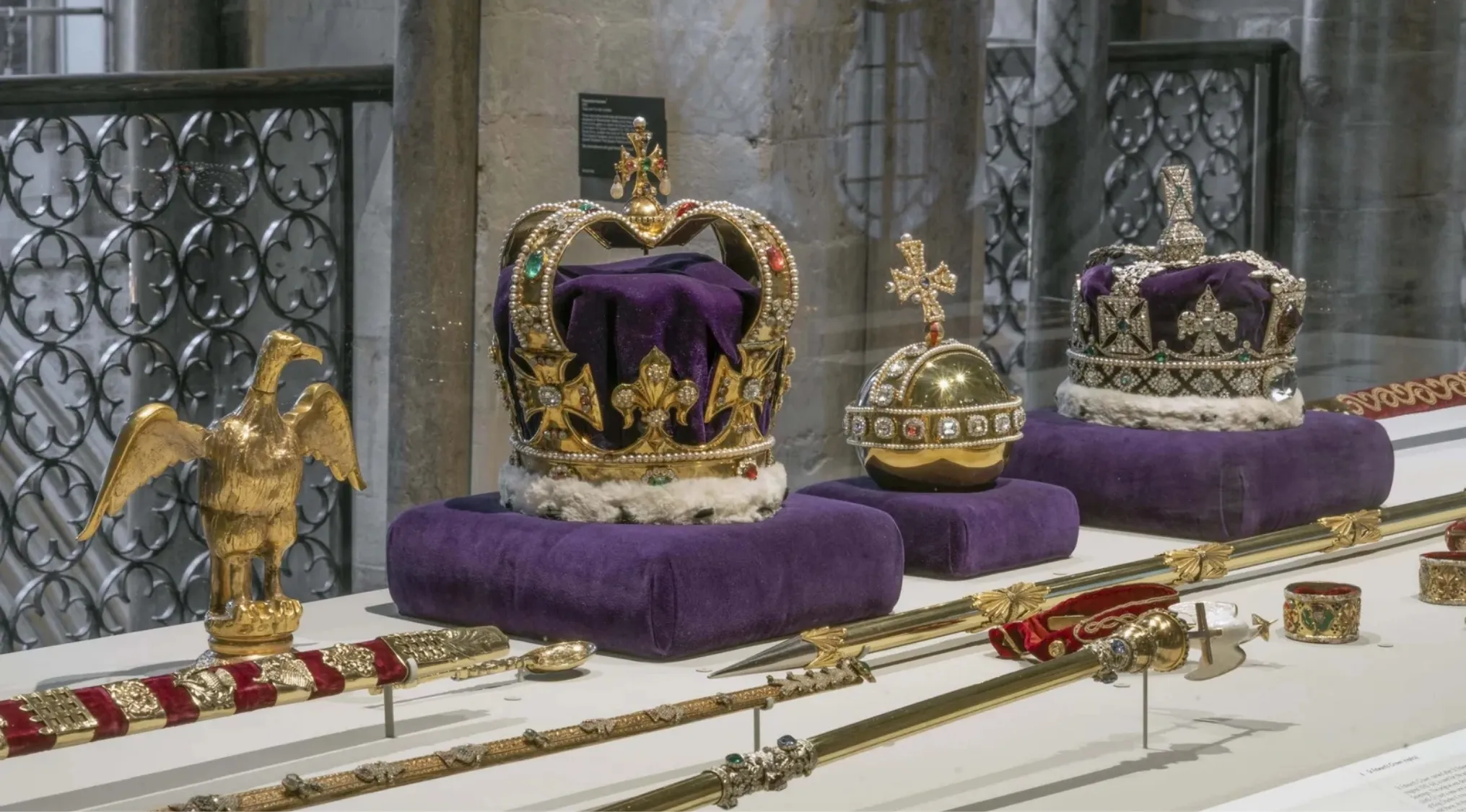 The Crown Jewels displayed on purple cushions with gold and jewel-encrusted crowns and royal scepters in a museum setting.