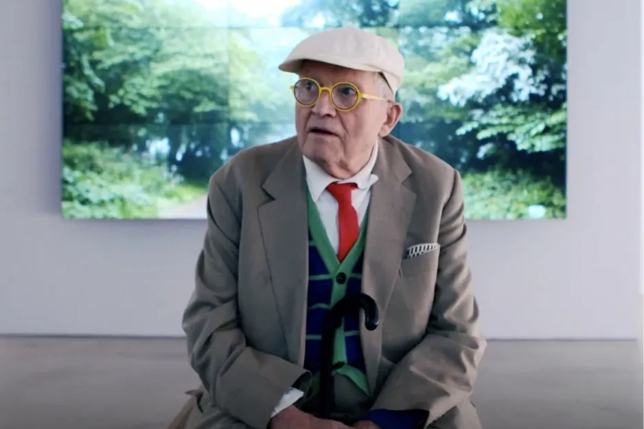 Elderly man wearing a hat and yellow glasses, seated indoors with artwork and greenery in the background, representing David Hockney.