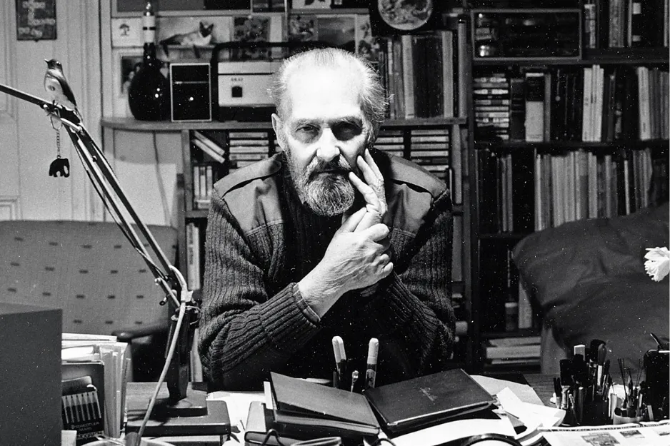 Black and white photo of an older man in a sweater at a cluttered desk with books and art supplies, Pop Art contributor.