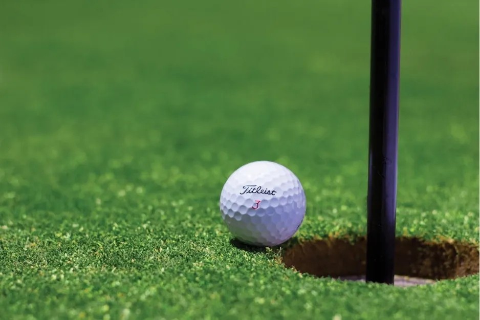 Golf ball near the hole on a green golf course with a flagpole casting a shadow on the grass.