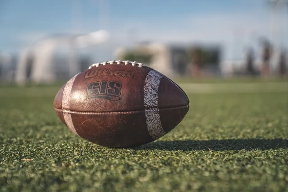 Rugby ball on green field with blurred players and stadium in the background under clear blue sky.