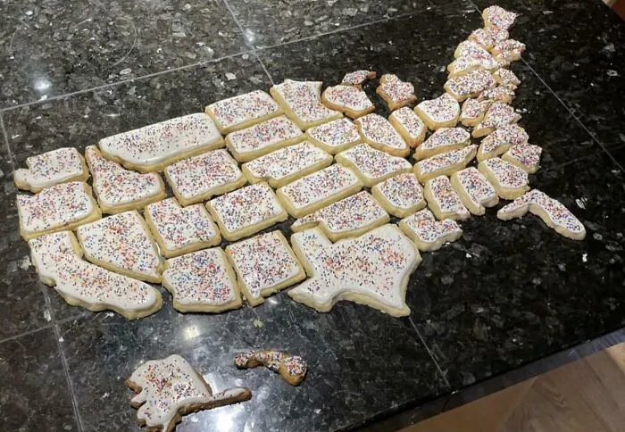 United States shaped cookies with white icing and colorful sprinkles arranged on a black countertop geography humor.