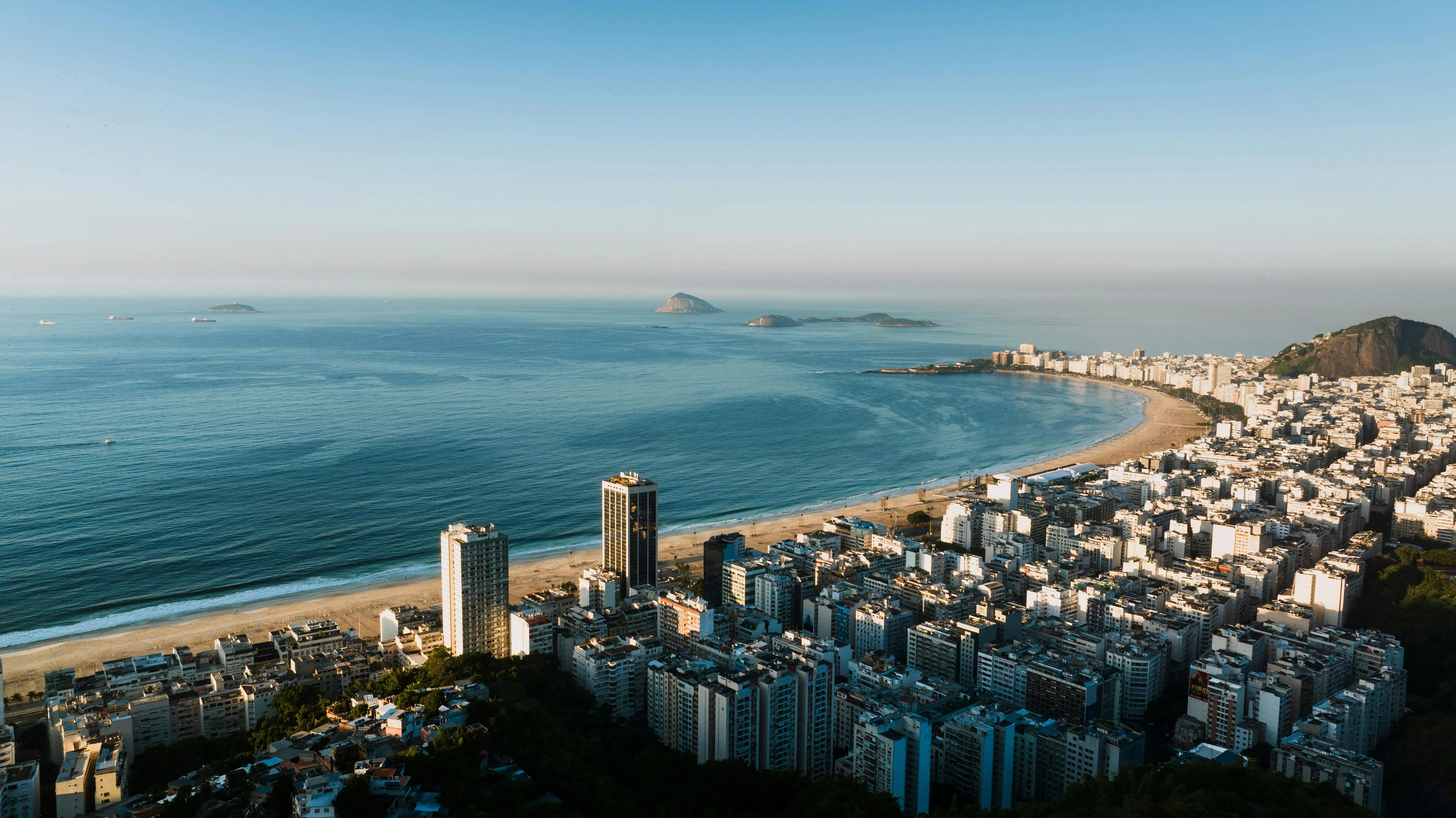 Coastal cityscape with mild climate view showing ocean, sandy shore, and buildings under clear blue sky at sunrise.