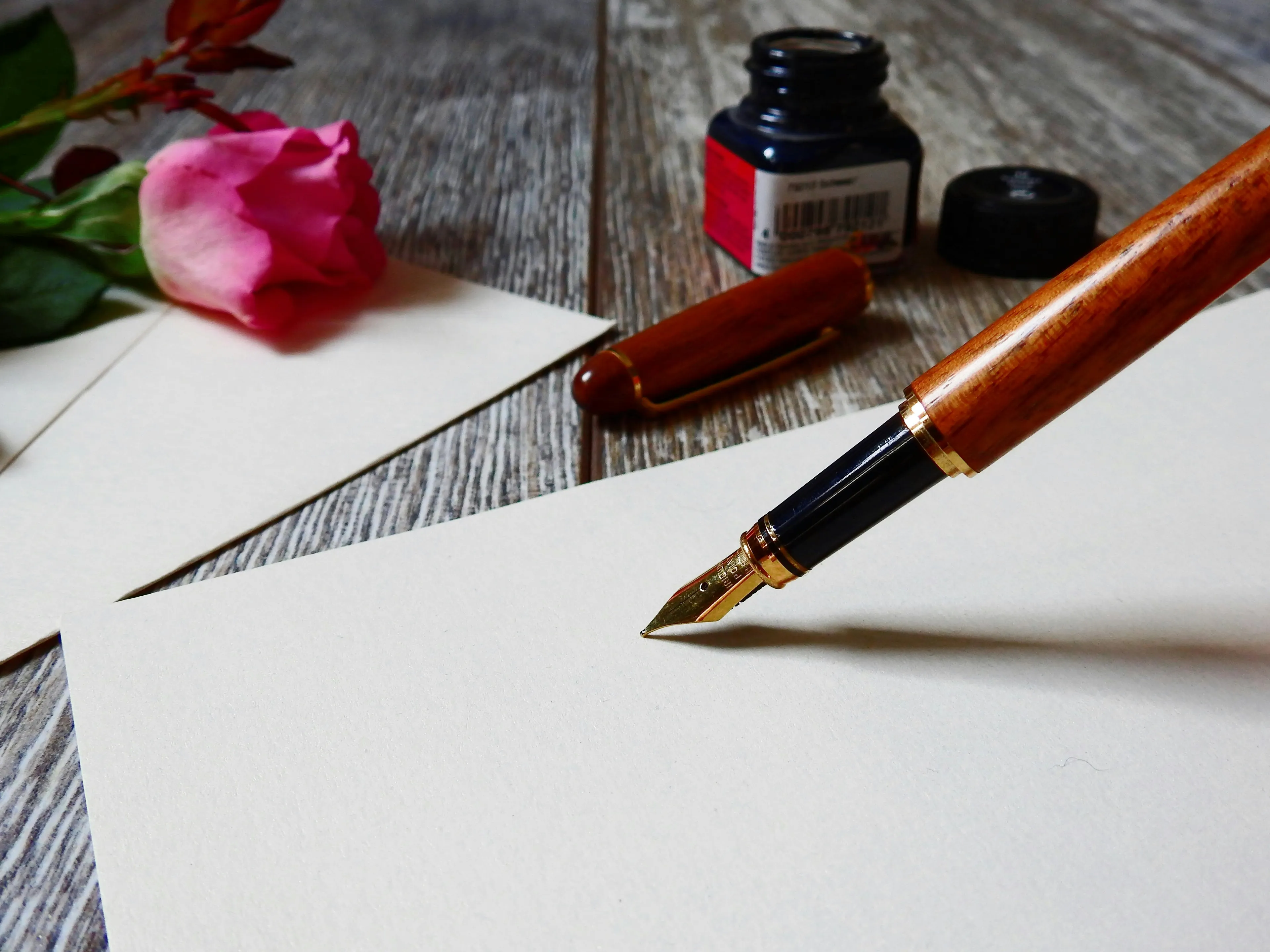 Fountain pen poised on blank paper with pink rose and ink bottle in the background for language grammar test.