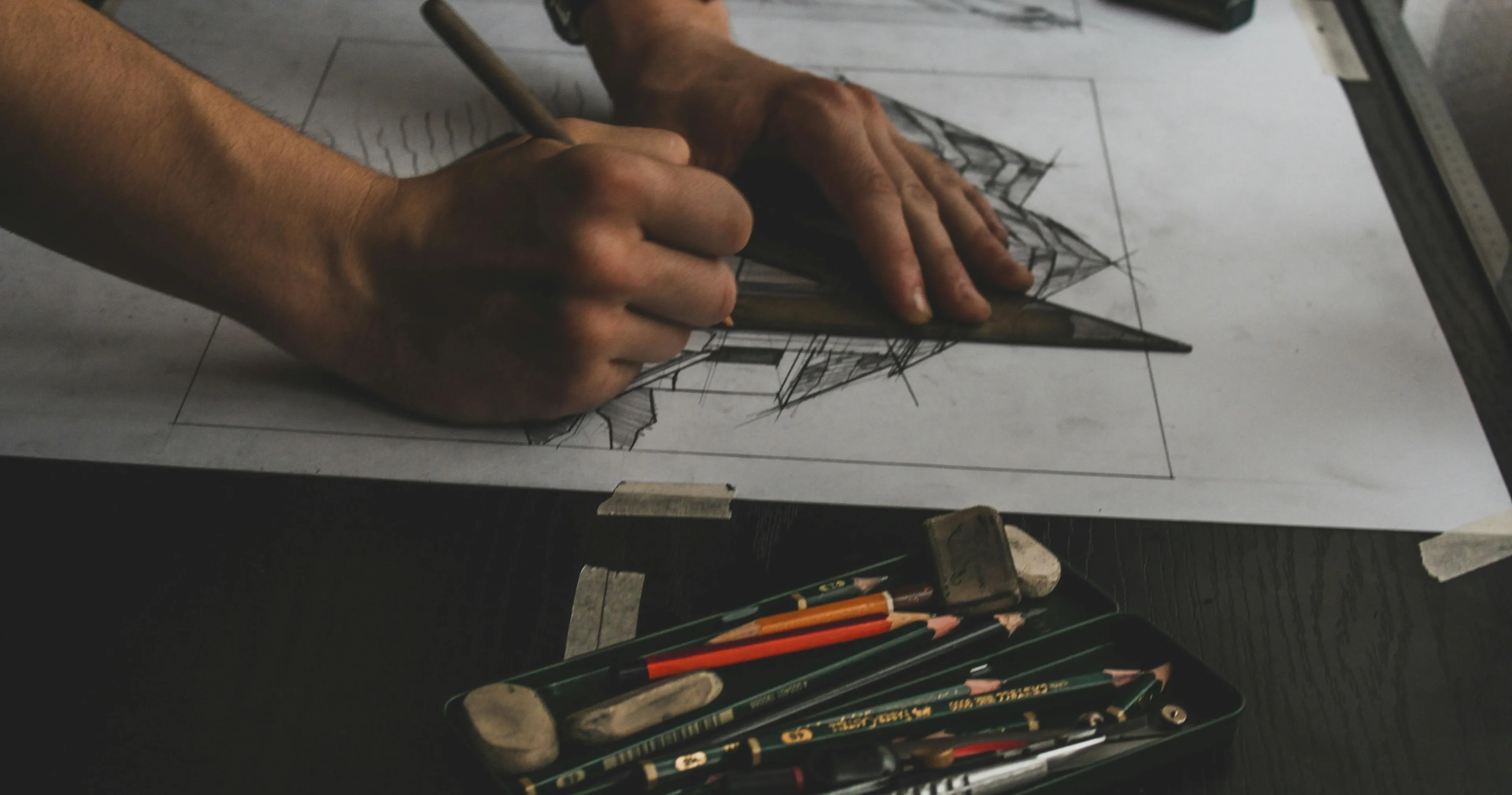 Hands drawing architectural perspective using pencils and a triangle ruler with drawing tools nearby on dark table.