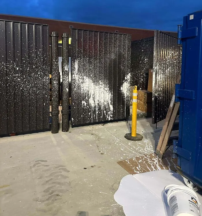White paint spilled and splattered on a warehouse floor, metal fences, and a yellow bollard in a funny photos moment.