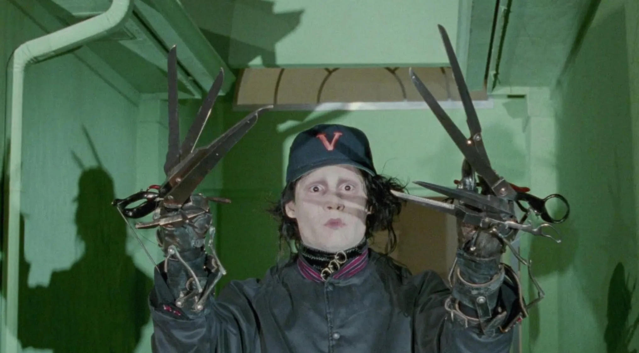 Edward Scissorhands character with scissor hands wearing a dark coat and cap inside a green room.