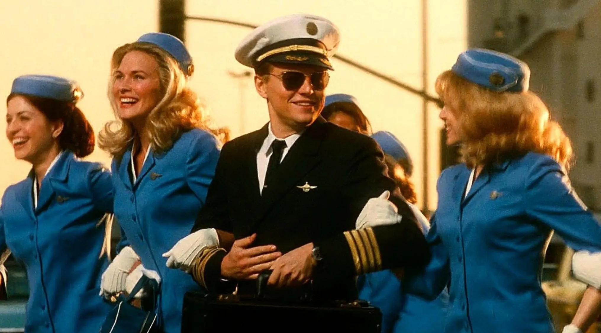 Leonardo DiCaprio in pilot uniform smiling with four women dressed as flight attendants in blue outfits from Catch Me if You Can movie.