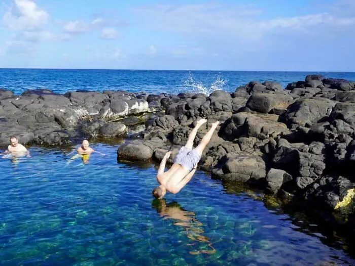 Man mid-flip into water near rocky shore with others swimming, capturing an unexpected better photo moment.