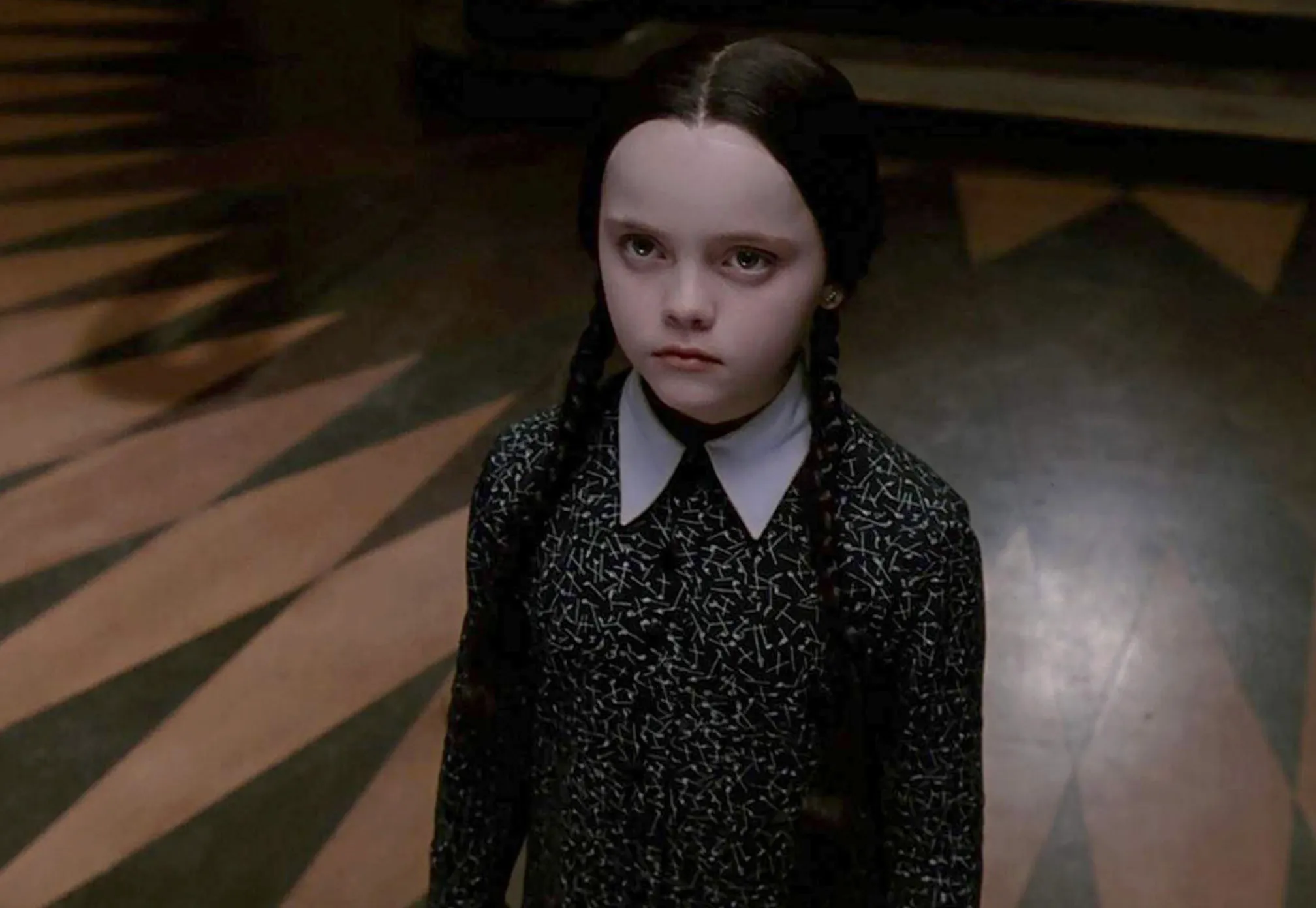 Wednesday Addams with braided hair and dark clothing standing on a patterned floor at Nevermore Academy.