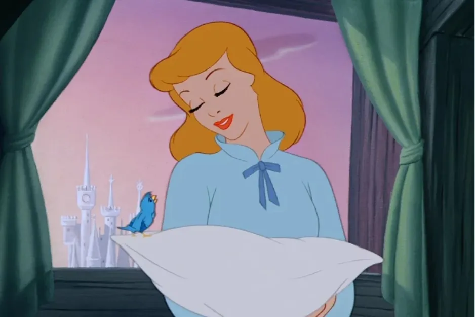 Cinderella smiling while holding a pillow with a blue bird on it, castle visible through window in the background.