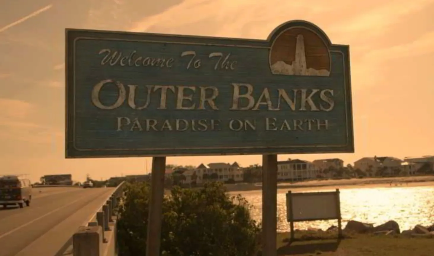 Welcome sign for Outer Banks, the fictional island setting of the TV show, with houses and water in the background.
