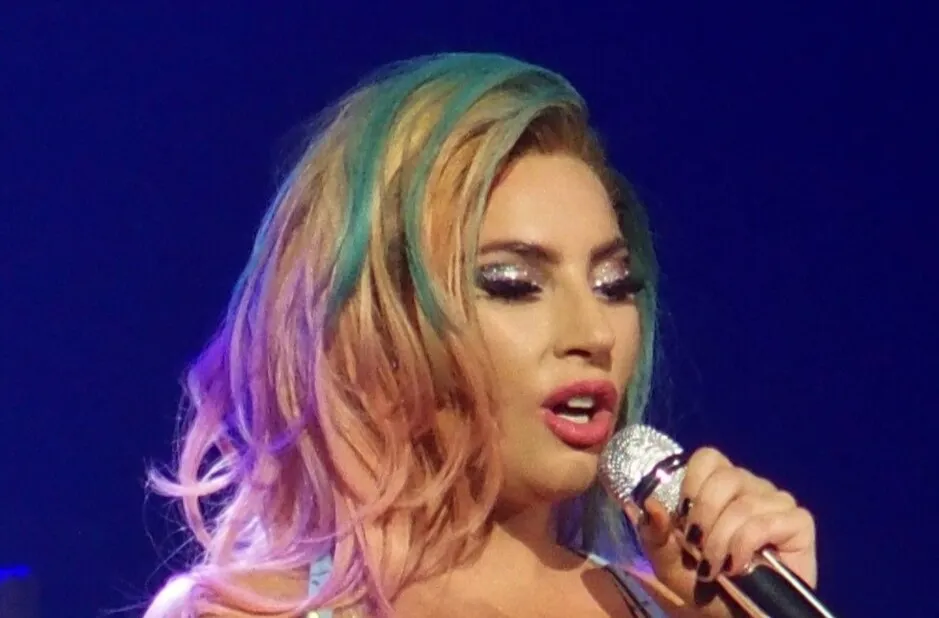 Lady Gaga performing at a concert with glittery makeup and colorful hair, holding a sparkly microphone.
