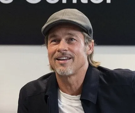 Brad Pitt wearing a gray cap and black jacket, smiling during an indoor event.